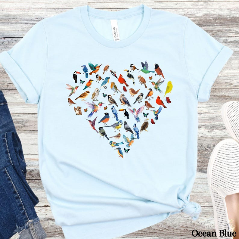 May include: A light blue t-shirt featuring a heart-shaped design made up of colourful birds. The birds are all different species and are arranged in a random pattern. The shirt is a casual style with short sleeves and a crew neck.