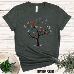 May include: A dark green t-shirt with a graphic of a tree with many colorful birds on the branches. The text "HEATHER FOREST" is at the bottom of the shirt.