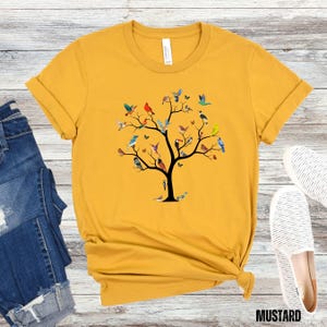 May include: A mustard yellow t-shirt with a graphic of a tree with many colorful birds on the branches. The tree is in the center of the shirt.
