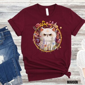 May include: A maroon t-shirt with a white cat surrounded by colorful flowers in a gold circle. The cat has a grumpy expression.