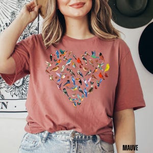 May include: A mauve t-shirt with a heart-shaped design featuring a variety of colourful birds. The birds are arranged in a heart shape, with the point of the heart at the bottom. The t-shirt is made of a soft, comfortable material and is perfect for everyday wear.