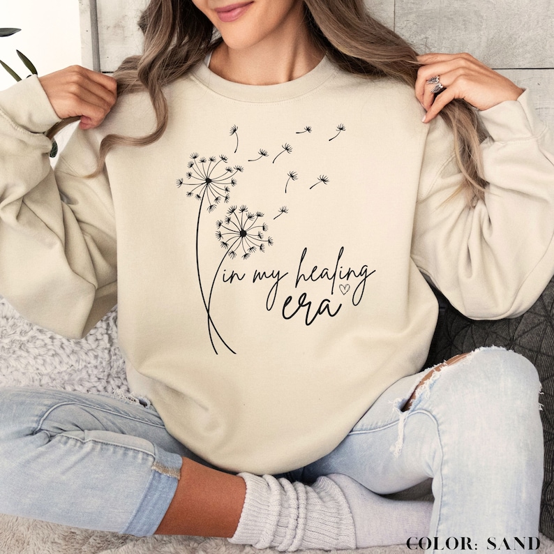 K&ouml;nnte beinhalten: Ein beiger Pullover mit einem Aufdruck von zwei L&ouml;wenzahnpflanzen und dem Text "in my healing era" mit einem kleinen Herzsymbol. Der Text ist in Schwarz.