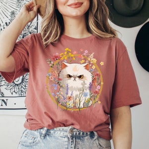 May include: A pink t-shirt with a white cat surrounded by colorful flowers. The cat has a grumpy expression and is sitting in a circle of flowers.