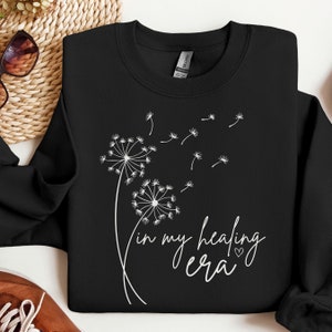 K&ouml;nnte beinhalten: Schwarzer Pullover mit einem wei&szlig;en L&ouml;wenzahnmuster und dem Text "in my healing era" mit einem kleinen Herzsymbol.