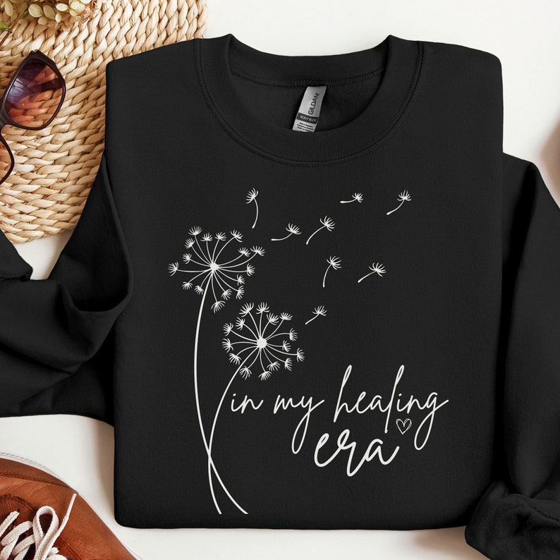 K&ouml;nnte beinhalten: Schwarzer Pullover mit einem wei&szlig;en L&ouml;wenzahnmuster und dem Text "in my healing era" mit einem kleinen Herzsymbol.