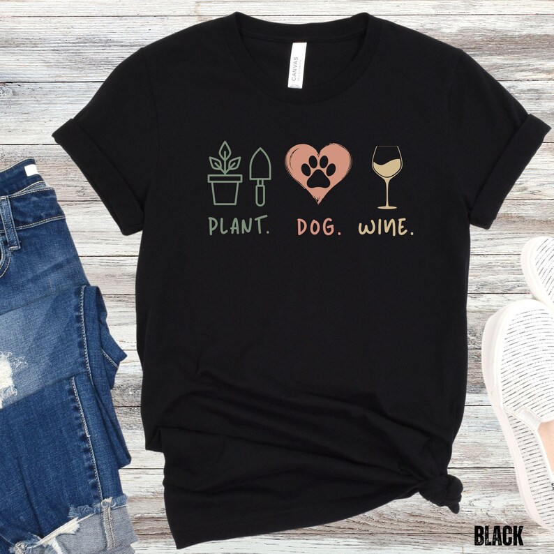 Pu&ograve; includere: Maglietta nera con un design grafico di una pianta, un cuore con un'impronta di zampa e un bicchiere di vino. Il testo "PLANT. DOG. WINE." &egrave; stampato sotto il grafico.