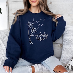 K&ouml;nnte beinhalten: Navyblauer Pullover mit einem wei&szlig;en L&ouml;wenzahnmuster und dem Text "in my healing era" mit einem kleinen Herz.