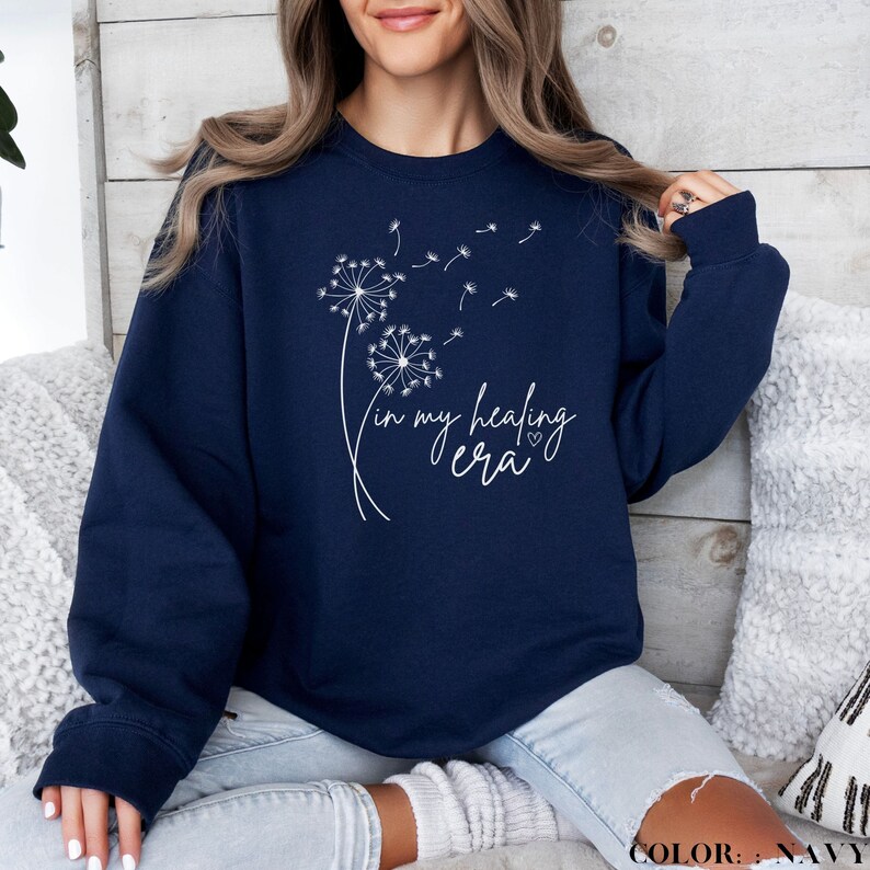 K&ouml;nnte beinhalten: Navyblauer Pullover mit einem wei&szlig;en L&ouml;wenzahnmuster und dem Text "in my healing era" mit einem kleinen Herz.