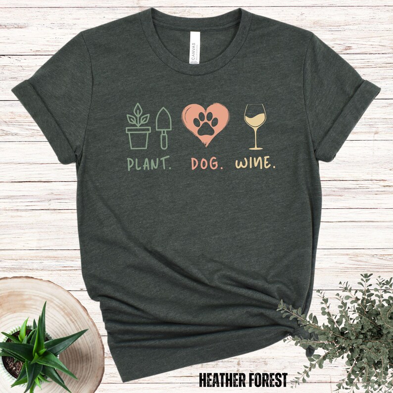 Pu&ograve; includere: Una maglietta verde scuro con un design grafico di una pianta, un'impronta di zampa all'interno di un cuore e un bicchiere di vino. Il testo "PLANT. DOG. WINE." &egrave; stampato sotto il grafico.