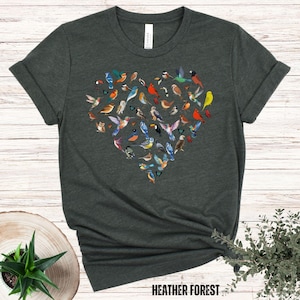 May include: A dark green t-shirt featuring a heart-shaped design made up of colourful birds. The text "HEATHER FOREST" is printed at the bottom of the shirt.