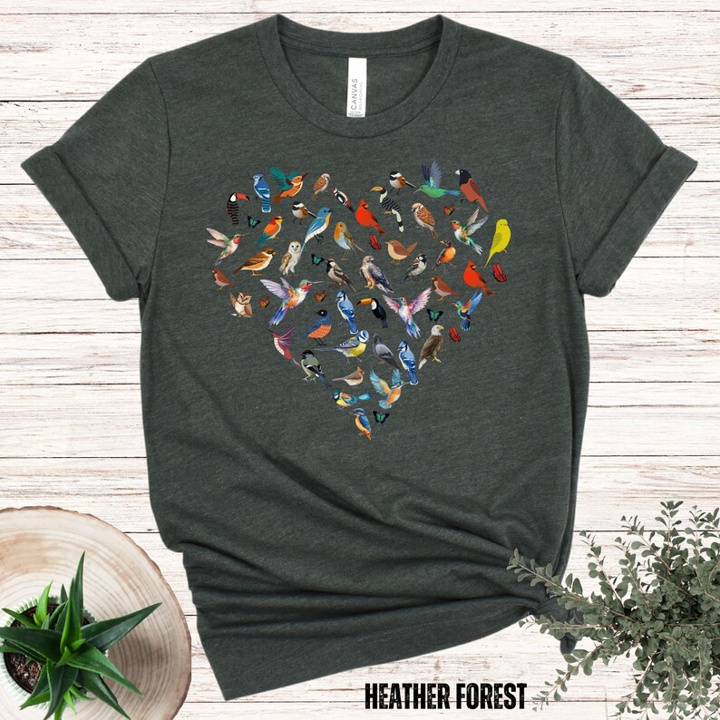 May include: A dark green t-shirt featuring a heart-shaped design made up of colourful birds. The text "HEATHER FOREST" is printed at the bottom of the shirt.