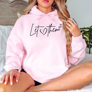 May include: Pink hoodie with a black graphic that says "Let them" with a heart in the middle. The hoodie has a drawstring hood and a kangaroo pocket.