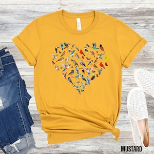 May include: A mustard yellow t-shirt featuring a heart-shaped design made up of colourful birds. The birds are arranged in a heart shape and are all different colours and sizes.