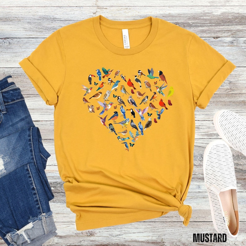 May include: A mustard yellow t-shirt featuring a heart-shaped design made up of colourful birds. The birds are arranged in a heart shape and are all different colours and sizes.