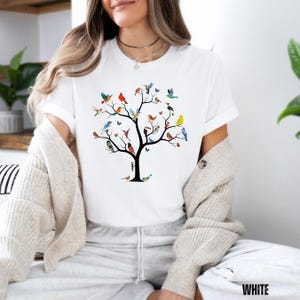May include: White t-shirt with a colorful graphic of a tree with birds on the branches. The tree is black and the birds are various colors, including blue, green, red, yellow, and orange.