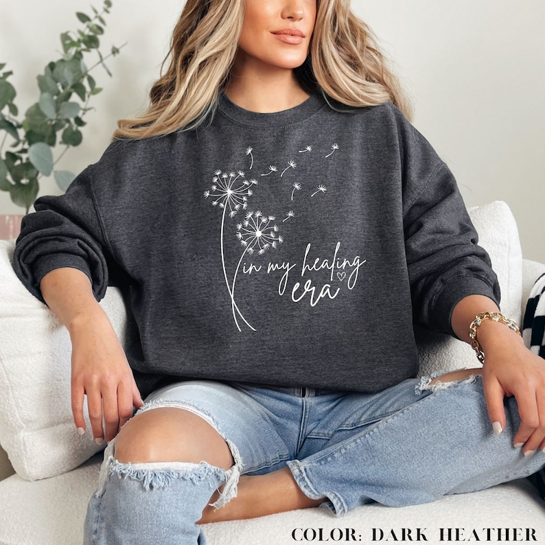 K&ouml;nnte beinhalten: Ein dunkel-heidesgraues Sweatshirt mit einem wei&szlig;en L&ouml;wenzahnmuster und dem Text "in my healing era".