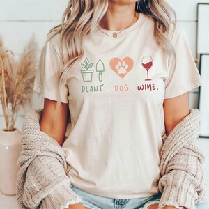 Pu&ograve; includere: Una maglietta color crema con un design grafico che presenta una pianta, un'impronta di zampa all'interno di un cuore e un bicchiere di vino. Il testo "PLANT. DOG. WINE." &egrave; stampato sotto il grafico.
