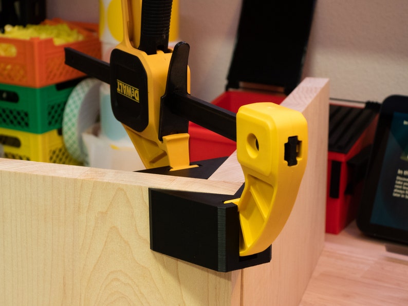 May include: A yellow and black corner clamp with a black base and a yellow lever. The clamp is holding two pieces of wood together.