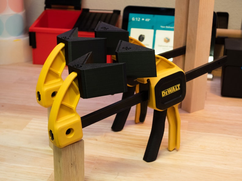May include: A yellow and black clamp with a black DEWALT logo is holding together three black 3D printed corner braces.