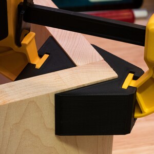 May include: A close-up of a black and yellow corner clamp holding two pieces of wood together. The clamp is made of plastic and has a metal screw.