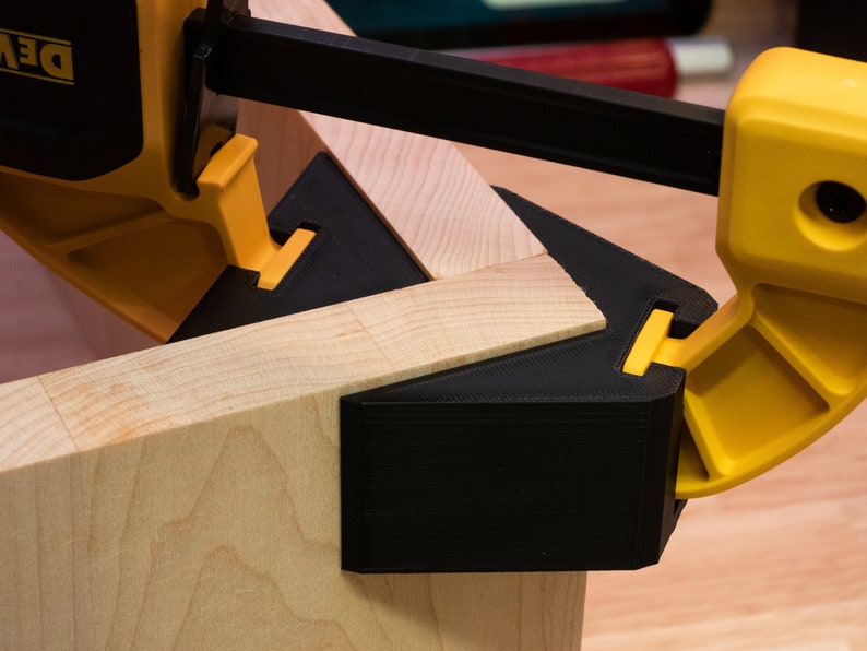 May include: A close-up of a black and yellow corner clamp holding two pieces of wood together. The clamp is made of plastic and has a metal screw.