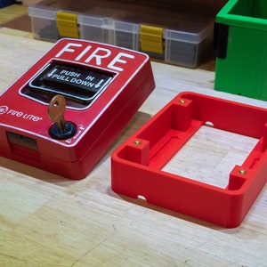 Könnte beinhalten: Ein roter Feuermelder mit einem Schlüssel im Schloss. Der Melder hat die Aufschrift "FIRE" und "PUSH IN PULL DOWN". Der Melder befindet sich neben einer roten Plastikbox mit vier Messing-Schrauben.