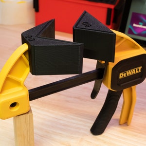 May include: A yellow and black DEWALT clamp with two black corner clamps, labeled "90" and "45", holding a piece of wood.