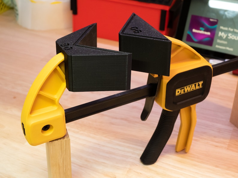 May include: A yellow and black DEWALT clamp with two black corner clamps, labeled "90" and "45", holding a piece of wood.