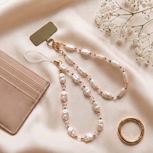 May include: A phone charm strap featuring irregular white faux pearl beads interspersed with small clear square beads and gold-toned metal spacers. The strap attaches to a gold-colored metal phone tab connector with a thin white cord loop.
