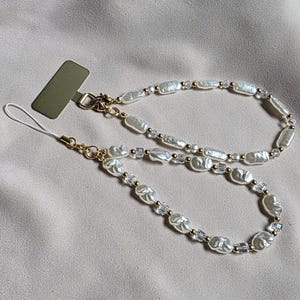 Puede incluir: Un charm para teléfono con un diseño de doble cadena. El charm está hecho de perlas blancas de forma irregular, cuentas de cristal transparente y detalles metálicos dorados. El charm se muestra sobre un fondo de tela de color neutro.