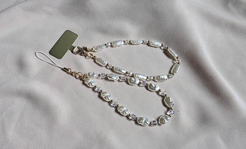 May include: A phone charm featuring a double strand design. The charm is made of white, irregularly shaped pearls, clear crystal beads, and gold-colored metal accents. The charm is shown on a neutral-colored fabric background.