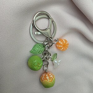 May include: A silver keychain with a lobster clasp, adorned with miniature fruit charms. The charms include green and orange citrus fruits, green leaves, and a small white charm. The keychain is set against a light pink fabric.