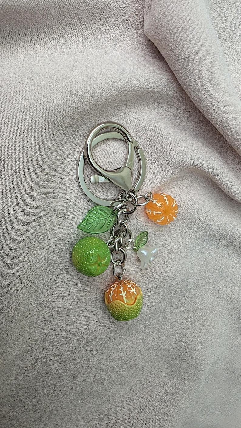 May include: A silver keychain with a lobster clasp, adorned with miniature fruit charms. The charms include green and orange citrus fruits, green leaves, and a small white charm. The keychain is set against a light pink fabric.