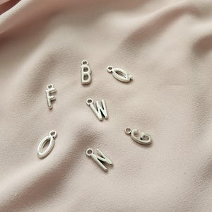 May include: Silver-colored metal alphabet charms, including the letters F, B, W, G, N, and O, are scattered on a soft, pink fabric. Each charm has a small loop for attaching to jewelry or other crafts. The letters are in a simple, sans-serif font.