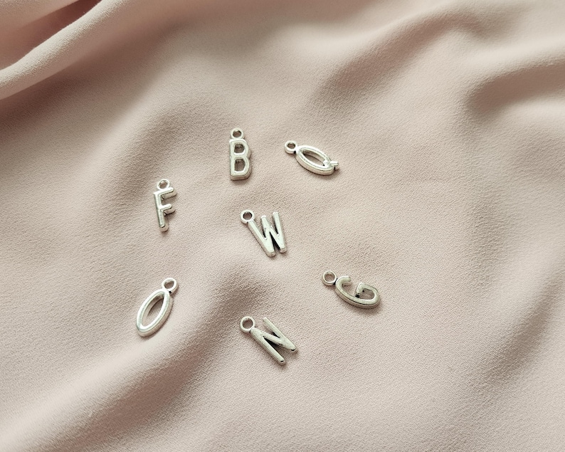 May include: Silver-colored metal alphabet charms, including the letters F, B, W, G, N, and O, are scattered on a soft, pink fabric. Each charm has a small loop for attaching to jewelry or other crafts. The letters are in a simple, sans-serif font.
