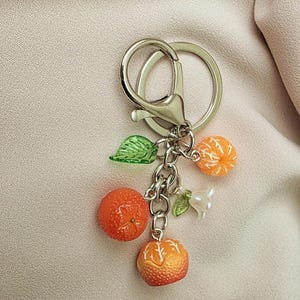 Może przedstawiać: Srebrny brelok do kluczy z karabińczykiem i dzielonym kółkiem. Zawieszka posiada trzy pomarańczowe zawieszki z owocami mandarynki, dwie zielone zawieszki z liśćmi i białą zawieszkę z perłą. Mandarynki są pomarańczowe i żółte.