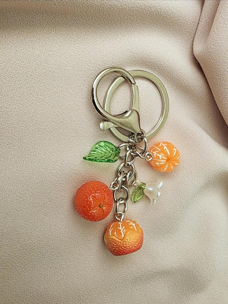 May include: A silver keychain with a lobster clasp and a split ring. The charm features three orange mandarin orange fruit charms, two green leaf charms, and a white pearl charm. The mandarin oranges are orange and yellow.