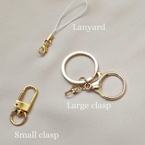 May include: A collection of gold-colored hardware accessories, including a small clasp, a lanyard with a white cord, and a large clasp, all displayed against a neutral background. The items are designed for attaching to keychains or other accessories.