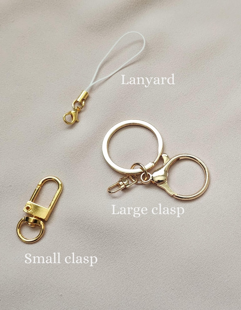 May include: A collection of gold-colored hardware accessories, including a small clasp, a lanyard with a white cord, and a large clasp, all displayed against a neutral background. The items are designed for attaching to keychains or other accessories.
