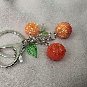May include: A silver keychain with a lobster clasp, featuring a cluster of orange fruit charms. The charms include three oranges, a white flower, and green leaves. The oranges have detailed textures.