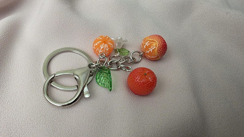 May include: A silver keychain with a lobster clasp, featuring a cluster of orange fruit charms. The charms include three oranges, a white flower, and green leaves. The oranges have detailed textures.
