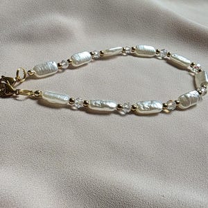 May include: A delicate bracelet featuring oblong, off-white pearls and small clear beads, strung together with gold-colored chain and accents. The bracelet is displayed on a neutral-toned fabric, with a gold-colored tag attached.