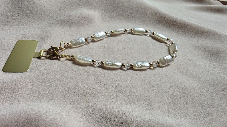 May include: A delicate bracelet featuring oblong, off-white pearls and small clear beads, strung together with gold-colored chain and accents. The bracelet is displayed on a neutral-toned fabric, with a gold-colored tag attached.