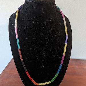May include: A multi-colored crocheted necklace with a rainbow pattern on a black display stand.