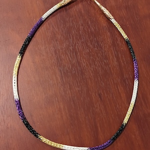 May include: A beaded necklace with a purple, white, and gold color scheme. The necklace is made of small beads that are woven together in a pattern. The necklace is closed with a gold clasp.