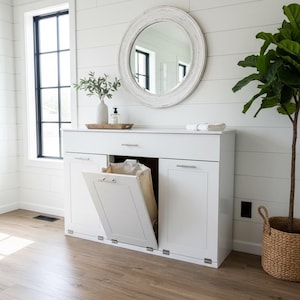 May include: A white cabinet with a laundry hamper, a round mirror, and a fiddle-leaf fig plant in a woven basket. The cabinet has a drawer and two doors, one of which is open, revealing a laundry bag. The room has white shiplap walls and wood flooring.