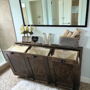 May include: A dark brown wooden laundry sorter with three compartments, each with a cream-colored fabric liner. The sorter has black handles and is placed in a bathroom setting. A black and white checkered basket sits on top.