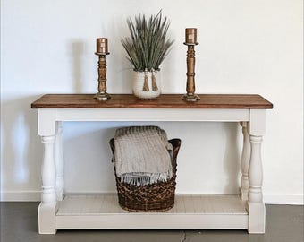 May include: A farmhouse-style console table with a stained wood top and white base. Two wooden candlesticks and a decorative plant in a white pot sit on the table. A woven basket with a gray blanket is below.