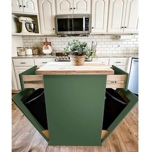 May include: A green kitchen island with a wooden countertop and two pull-out trash bins. The island has a dark green exterior and is set against a white kitchen with a microwave and cabinets. A plant in a woven basket sits on the countertop.