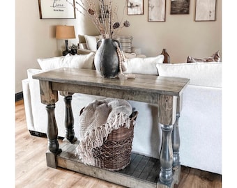 May include: A rustic wooden console table with a gray finish, featuring turned legs and a lower shelf. A woven basket with a fringed throw sits beneath the table. A dark vase with decorative branches and artwork adorn the background.